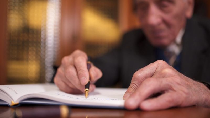 An elderly man writes a legal letter with a pen on a notebook at a wooden desk