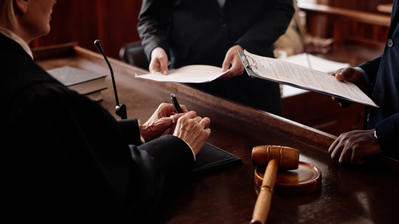 A judge receives legal documents from two individuals inside a courtroom