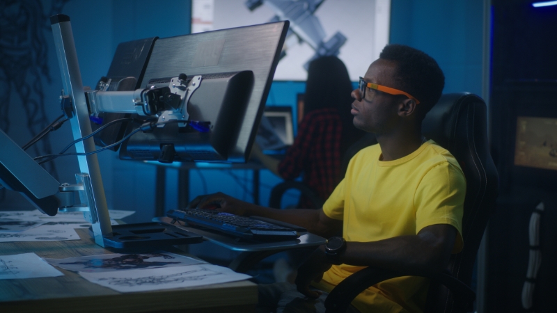 A game developer in a yellow shirt works at a dual-monitor setup in a modern studio