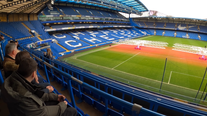 Fans sit inside a large European stadium with blue seats and a clear view of the pitch