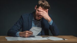 Man holding paperwork at a desk with his hand covering his face, looking overwhelmed