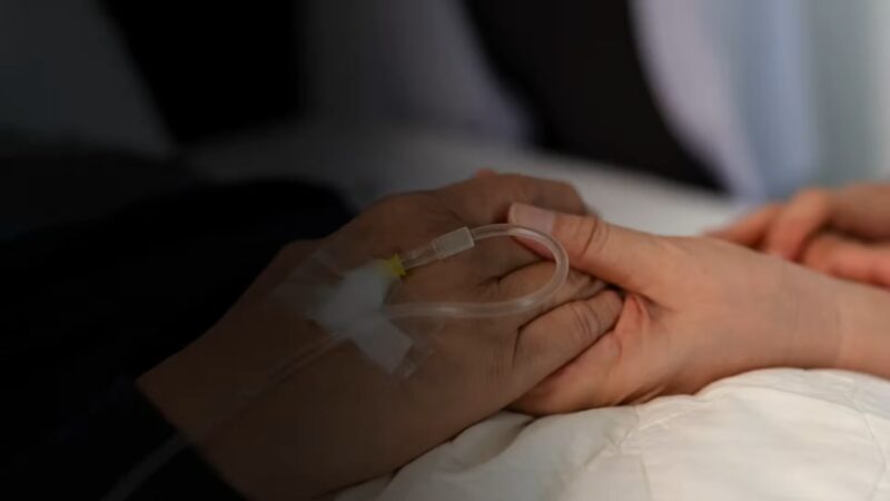 A patient with an IV holds another person’s hand in a hospital bed.